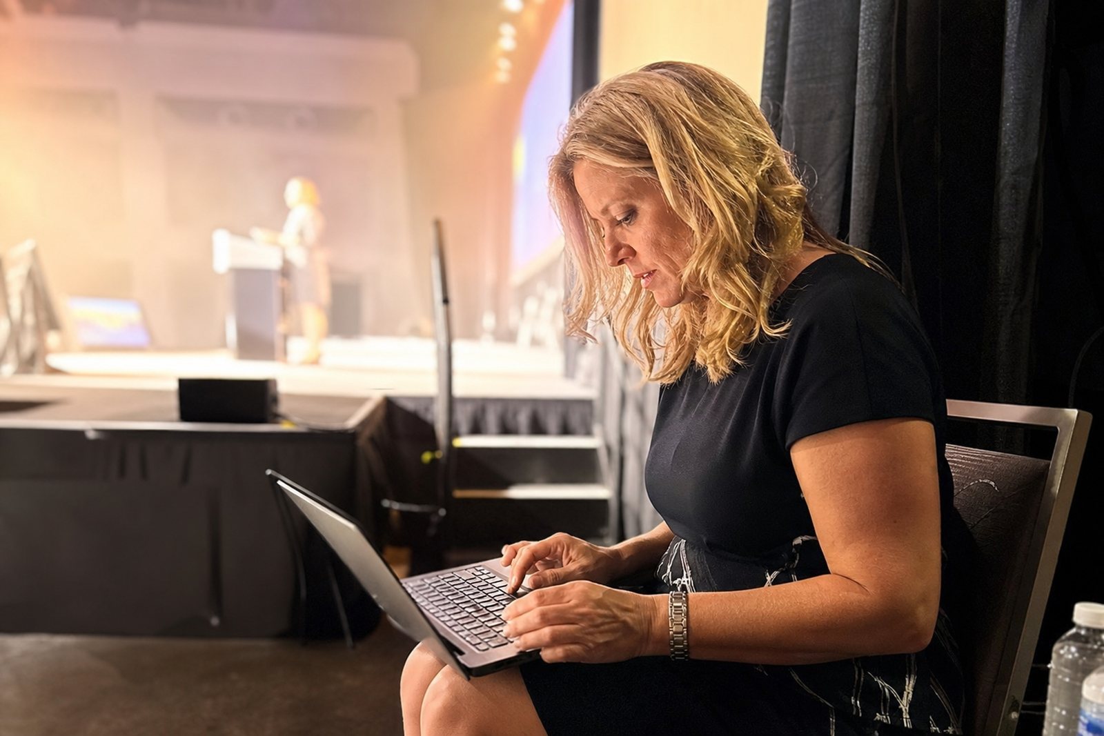A professional speaker reviews her notes on a laptop backstage beside a lit conference stage