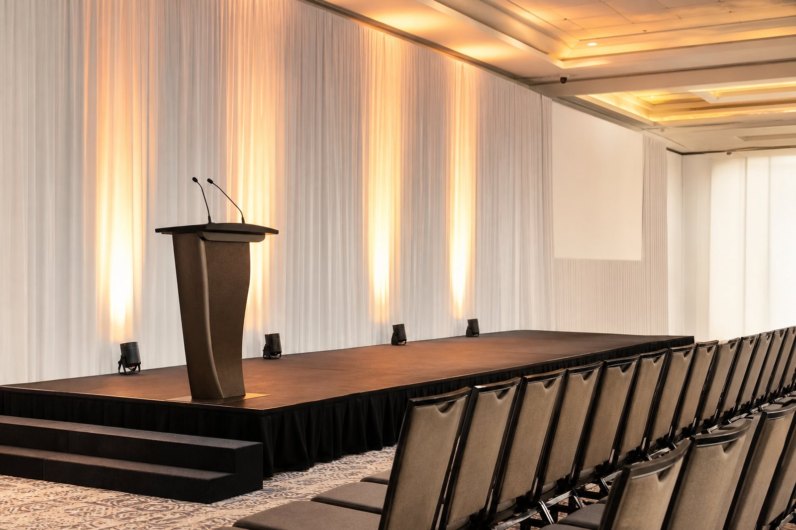 An empty conference stage with a lectern, warm spotlights and rows of chairs in a bright event hall